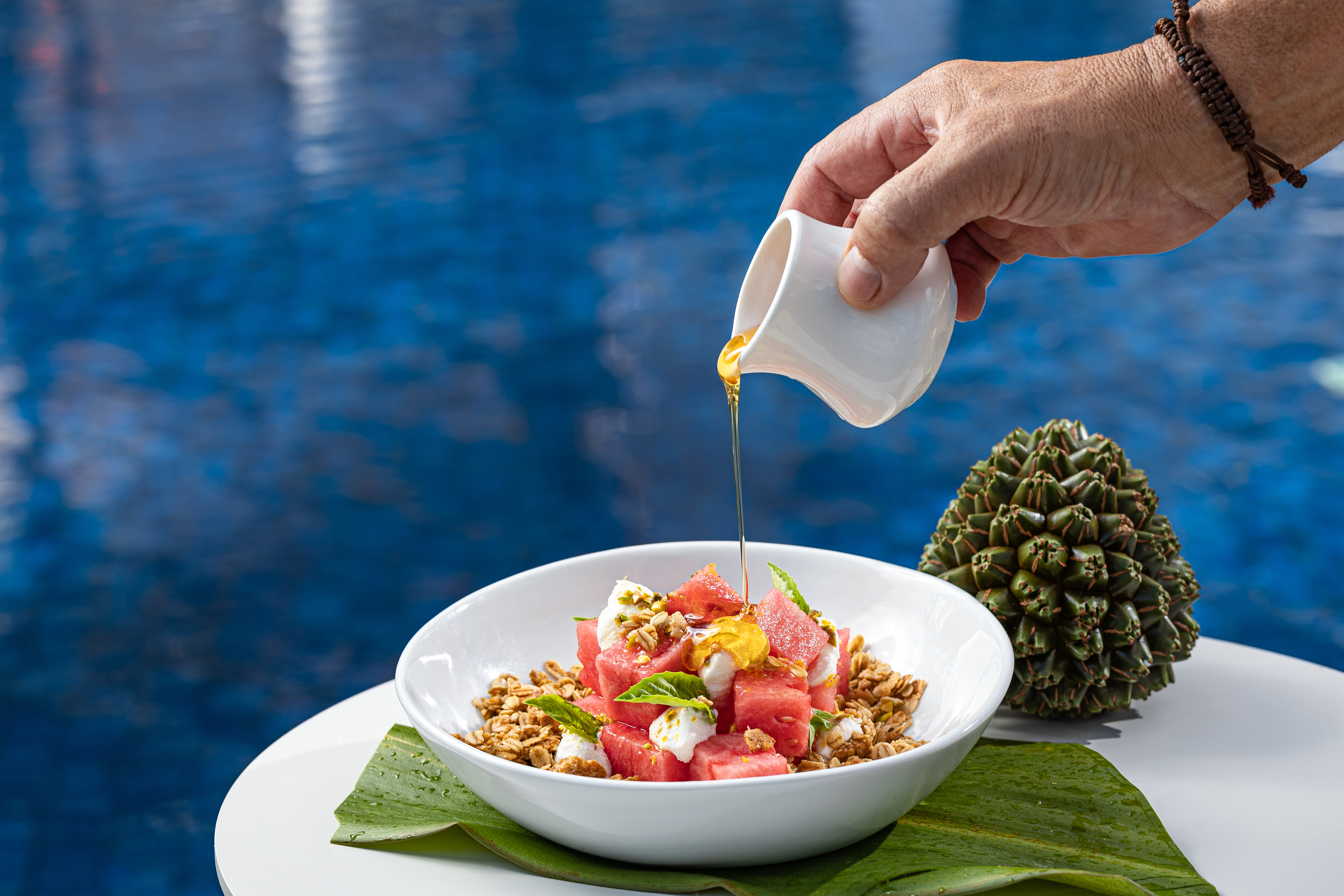 Hand pouring honey over a breakfast bowl with watermelon, granola, cheese, and mint, with a pool in the background.