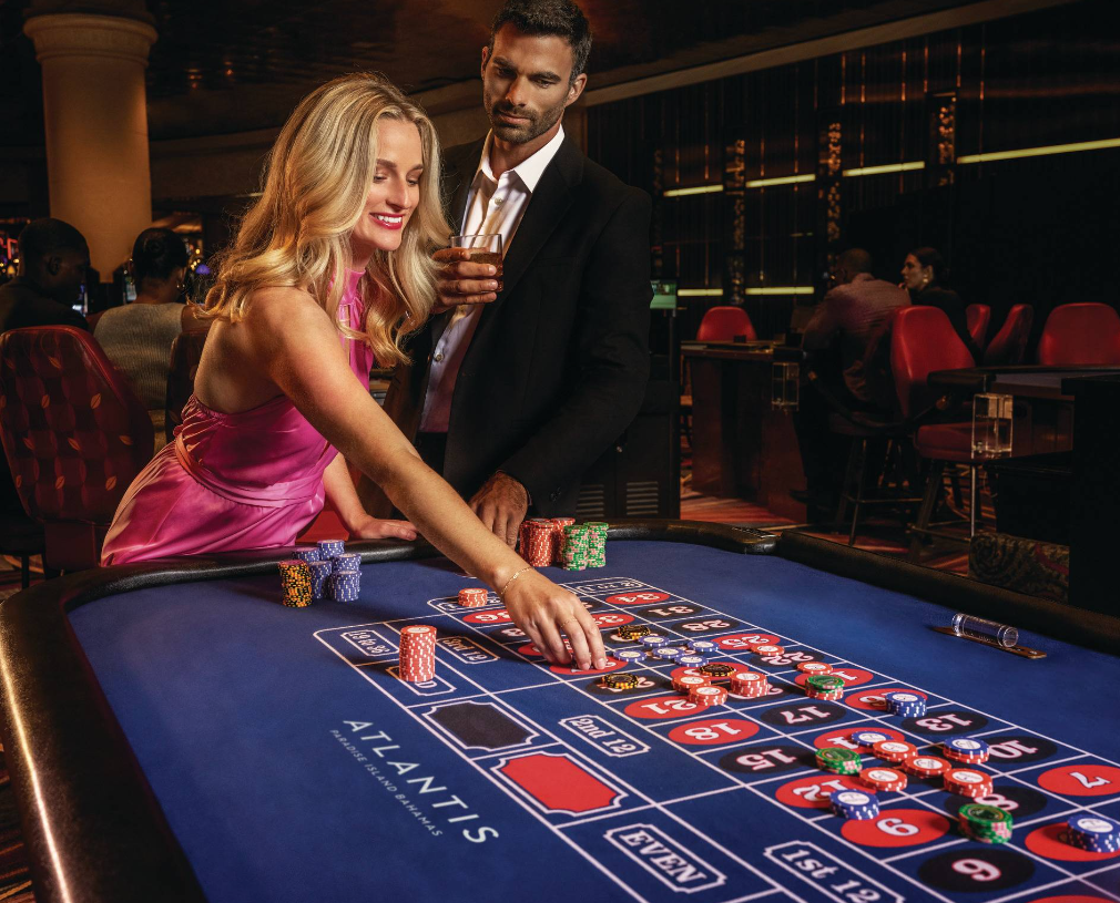 A woman in a pink dress places a bet at a roulette table while a man stands beside her with a cocktail.