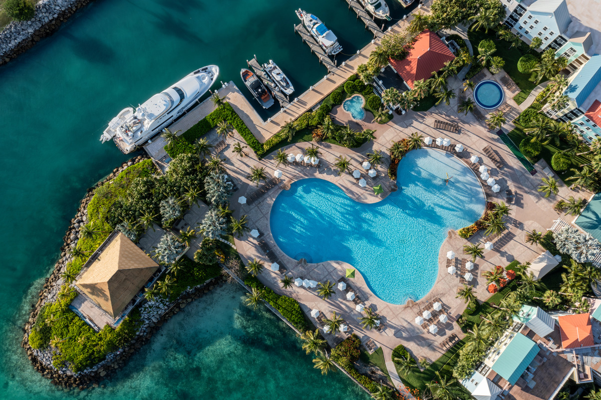 Aerial view of a resort pool framed by palm trees and lounge chairs with a marina full of yachts and boats nearby.